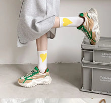 Load image into Gallery viewer, Person wearing white socks with yellow hearts and green sneakers, sitting on a gray crate.